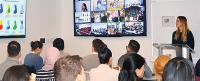 A presenter stands at the front of a classroom while students watch multiple screens showing video calls and presentations.